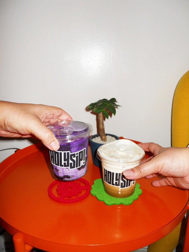 Two hands holding vibrantly colored Holy Sip drinks on an orange table.